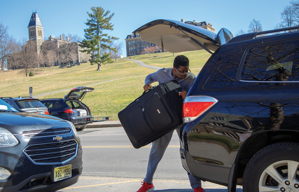 A student loads a suitcase into the back of an SUV on a sunny day. In the background, libe slope and the clocktower.