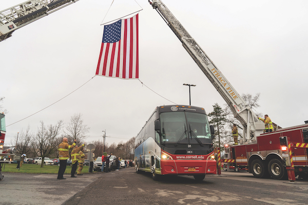 A firetruck extends its ladder and hangs an American flag over the road. a Cornell bus drives beneath it, while a crowd of people clap and cheer.